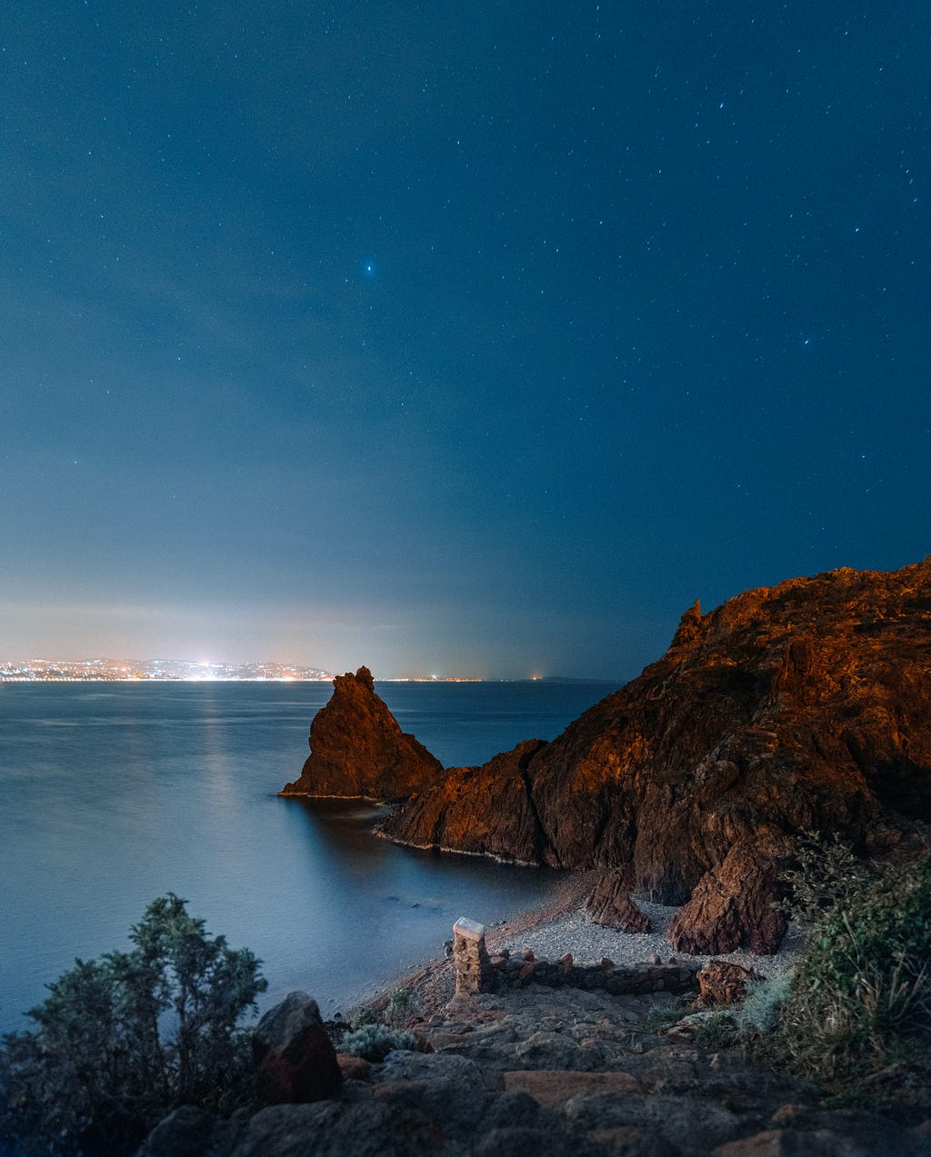 This is a picture of the sea and sky at night. The water and sky are both dark blue. We can also see stars in the sky. There is also a rock above the sea. Stonerock is dark brown.