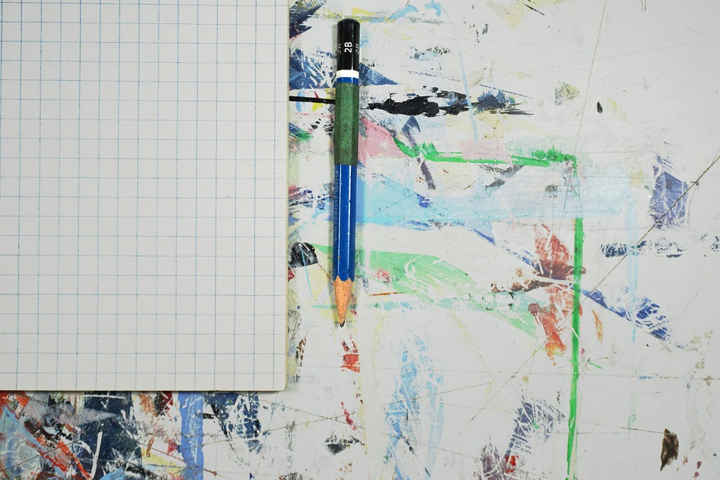 A blank grid notebook laying open on a paint-splattered desk with a pencil next to it.