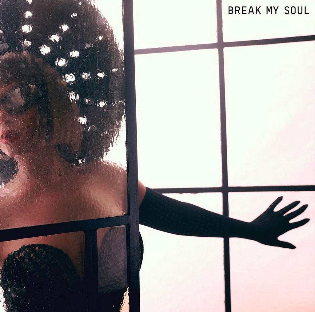 Cover art for “Break My Soul,” featuring Beyoncé posing with her arms spread open, standing behind frosted glass. She is wearing a black top, black full length gloves, a large black hat and black sunglasses.