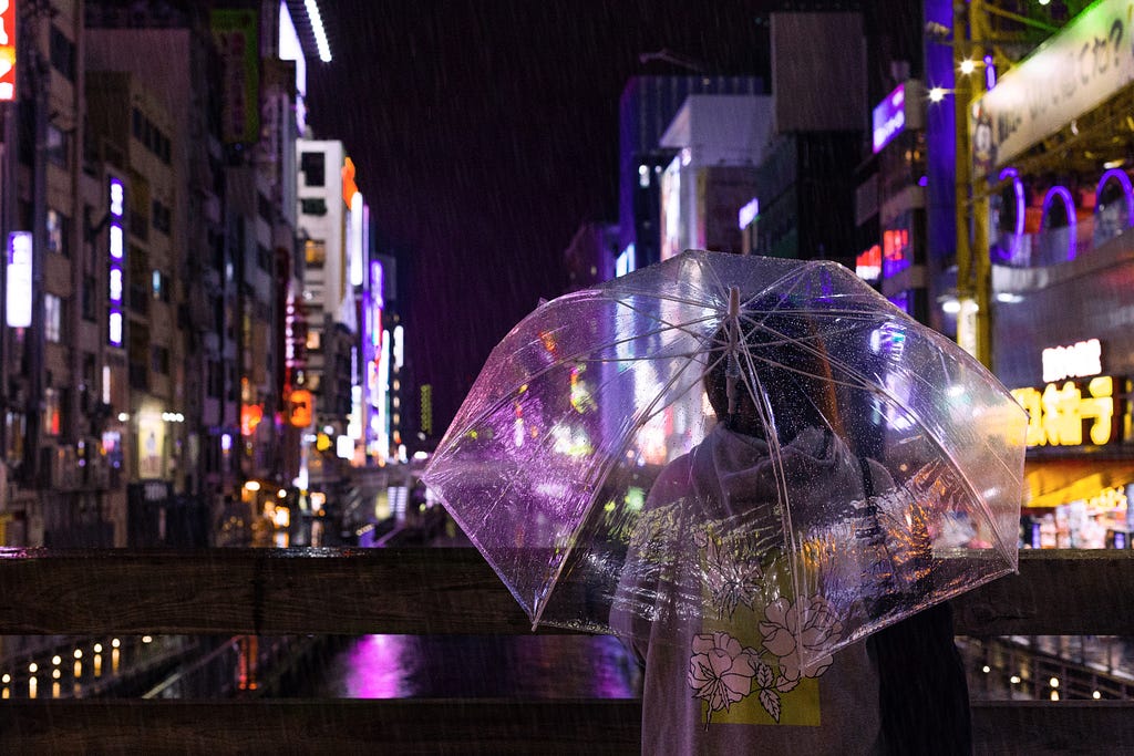 Person standing in city street with clear umbrella. They are to the right of the frame with bright neon lights on the buildings. As you look down the street, each building is bright with neon lights, billboards, or other lights. The person is in a gray hoodie with green and white Hawaiian flowers on the back.