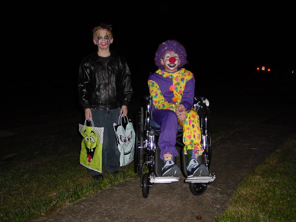 Two kids on Halloween ready to trick-or-treat. The little boy is dressed up as Eric Draven from the movie “The Crow” in all dark clothing. He is holding two trick or treat bags, one a green witch and the other a vampire. The little girl in a wheel chair is dressed up as a clown with purple hair and a bright red nose. The photo is from 2002.