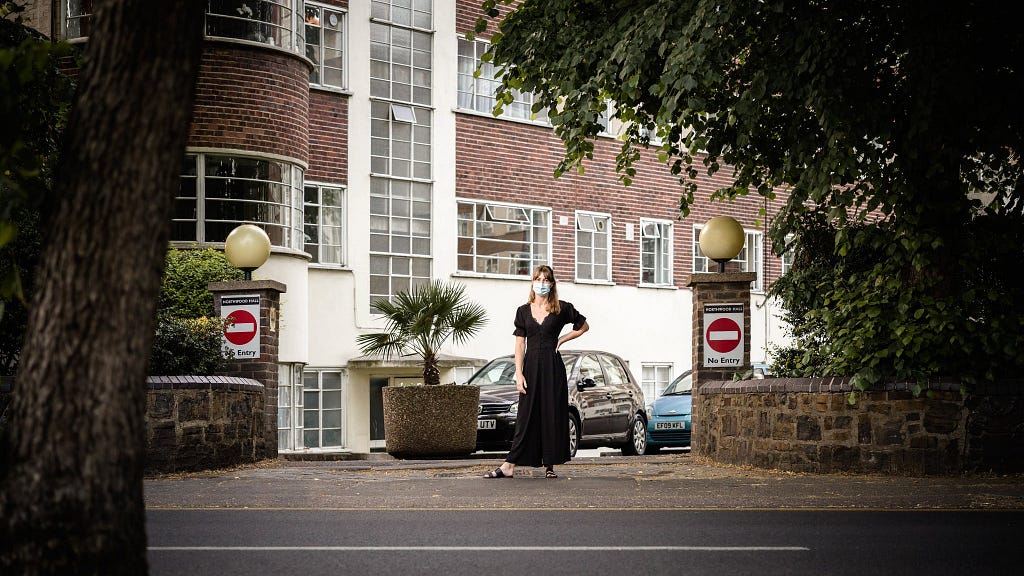 Photograph of a woman in a black dress and wearing a blue face-mask standing in the middle of an exit to a residential building. Two signs displaying the do not enter circle with white dash appear on pillars either side of the exit. The shot is captured from the other side of the road and features the trunk of a tree, the road and part of the pavement in the foreground.