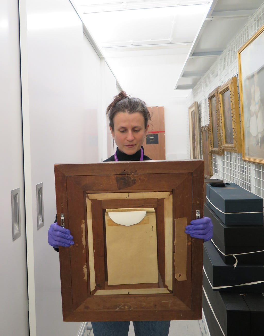 A conservator wearing purple nitrile gloves, carries a framed painting with one hand either side of the painting and the glazed side facing towards her