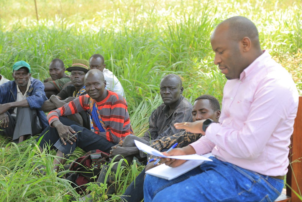 A man sitting outside in a chair with papers in his hand explains information to a group of men sitting on the ground among lush green grass.