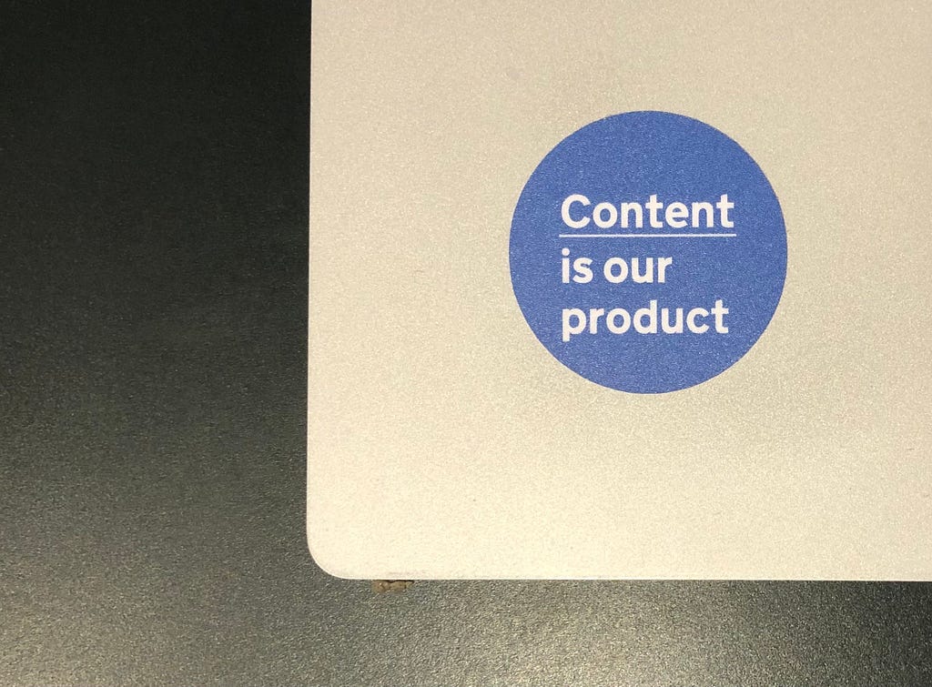 A blue sticker that says ‘Content is our product’ on a silver laptop