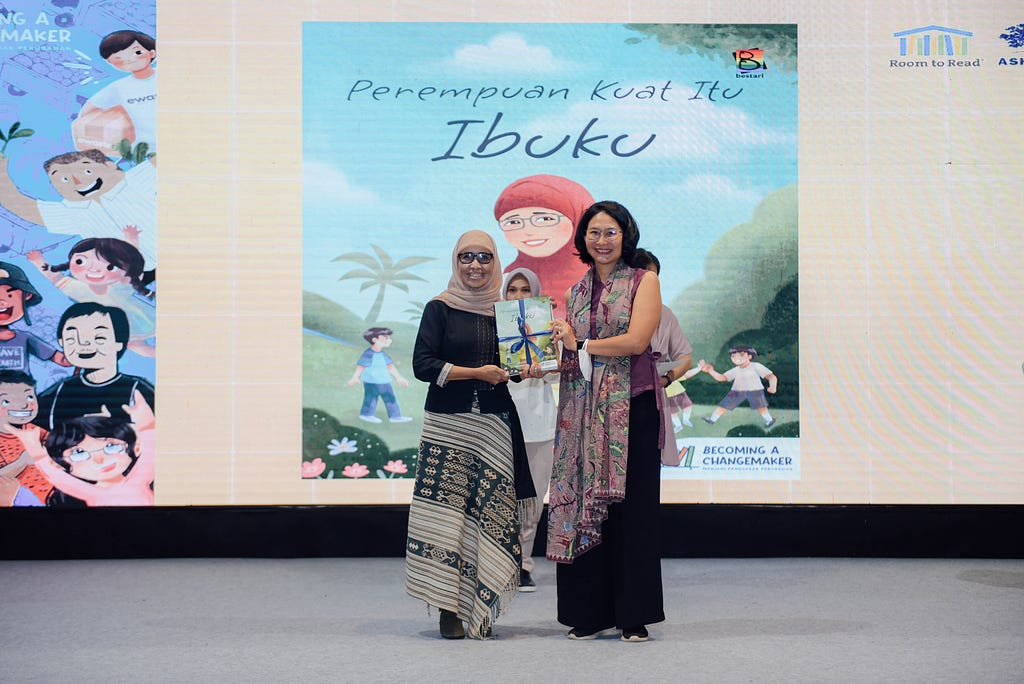 Two women pose in front on a large blue book image titled “Ibuku” holding a smaller version of the book. The woman on the left is wearing a pink hijab and patterned flowing skirt. The woman on the right is wearing glasses and a black dress with a colorful shawl draped over her shoulders.