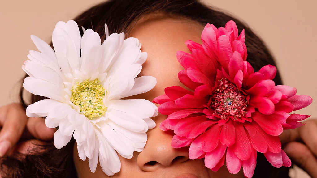 A black woman holds two flowers over her eyes. The right one is a white flower and the left one is a pink flower.