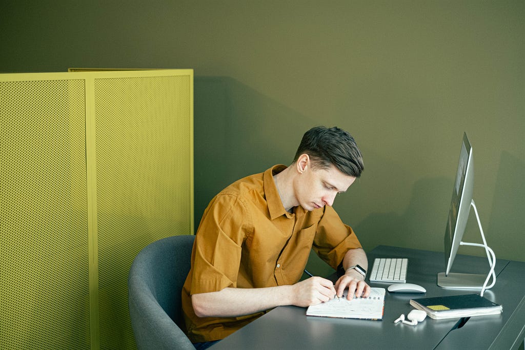 Man writing in a notebook, in front of a computer.