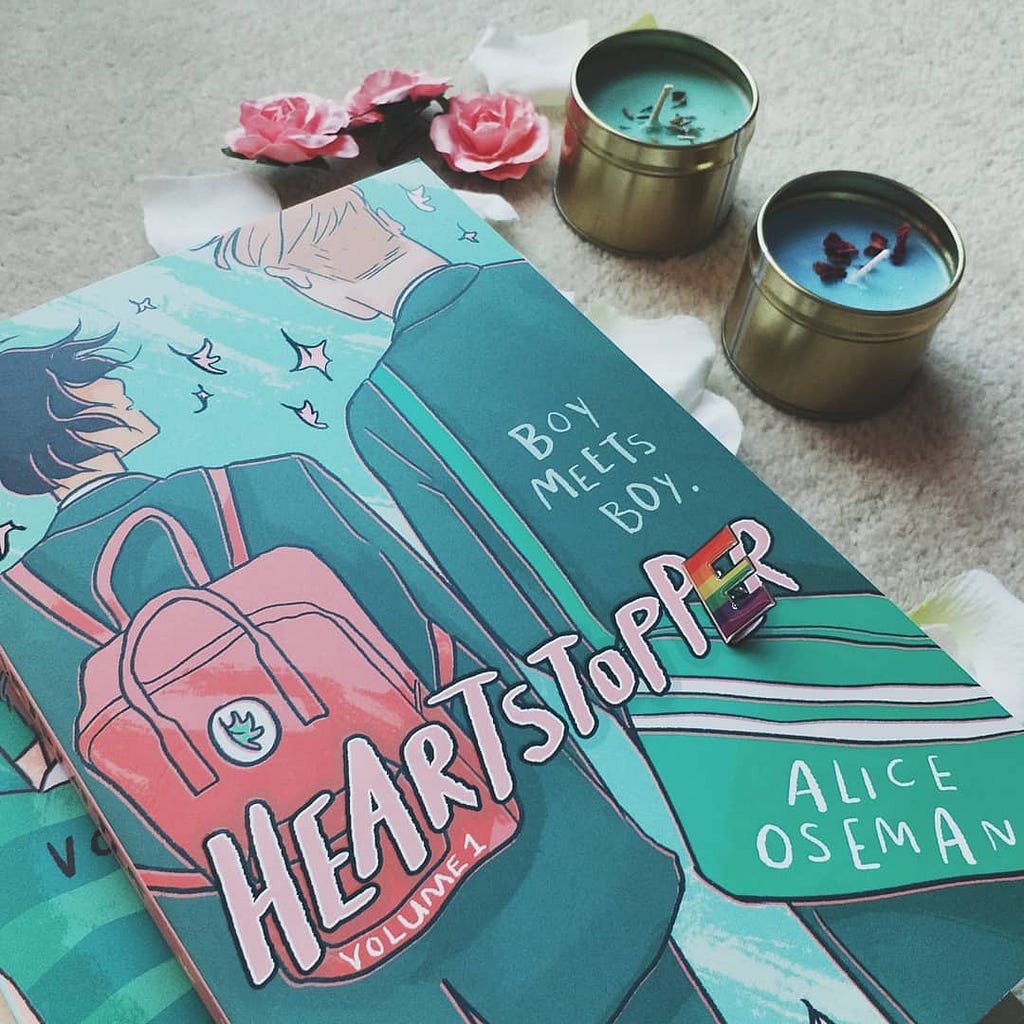 Volume 1 of Heartstopper by Alice Oseman, photographed with flowers, blue and green candles and a rainbow pin.