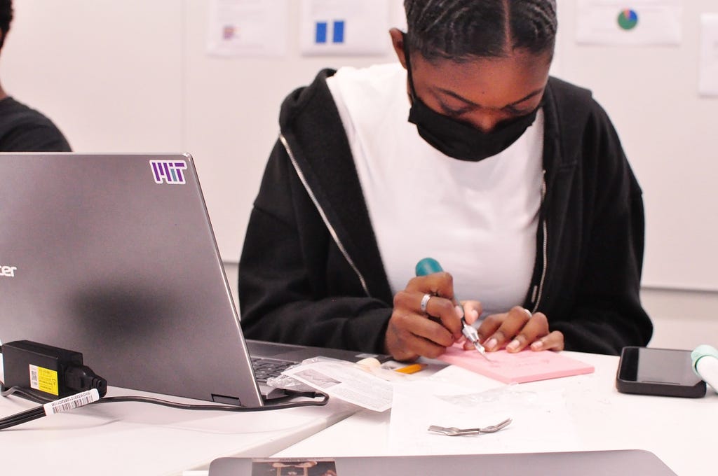 Photo of a teenaged girl wearing a mask and creating a rubber block stamp. A laptop is propped open beside her with an MIT sticker on it.