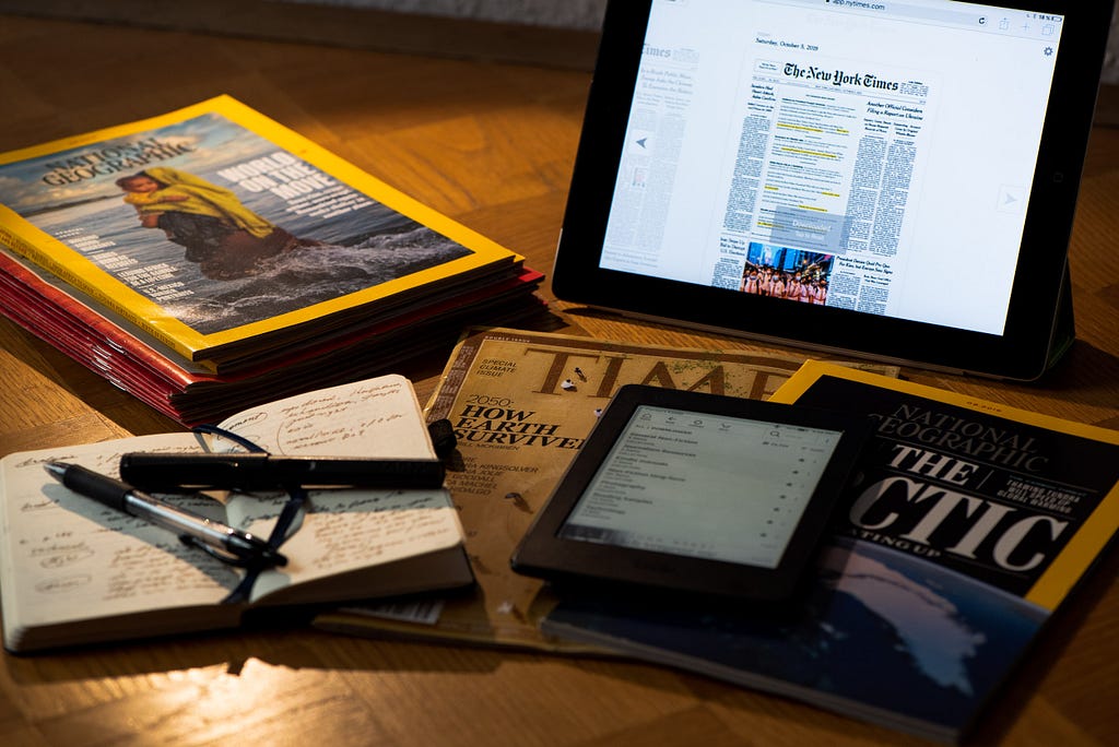 Display of magazines and newspapers, in print and digital, along with a Kindle and a pocket notebook and pens on a desk.