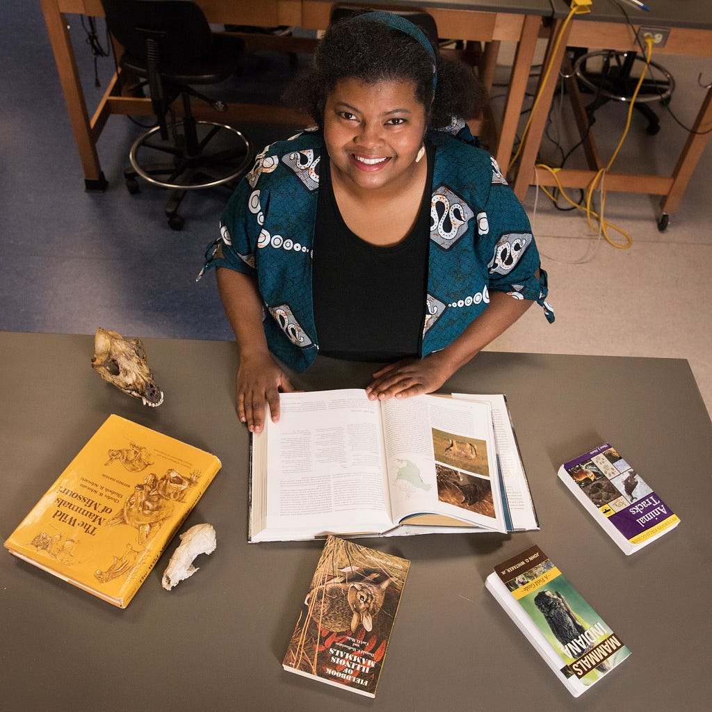 A person sitting at a table with several books. Visible book titles include &ldquo;The Hunters or the Hunted?&rdquo; by Robert W. Sussman, &ldquo;Primate Behavioral Ecology&rdquo; by Karen B. Strier, &ldquo;Primates in Perspective&rdquo; edited by Christina J. Campbell et al., and &ldquo;The Primate Anthology&rdquo; edited by Russell L. Ciochon and Richard A. Nisbett. An open book in front of the person contains text and images related to primates or anthropology.