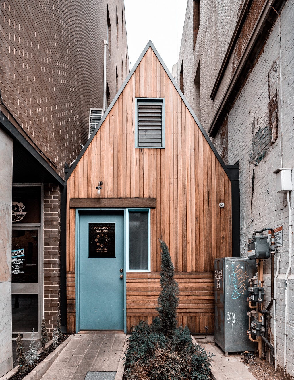 A tiny wooden house with a blue door, built between two brick building walls.