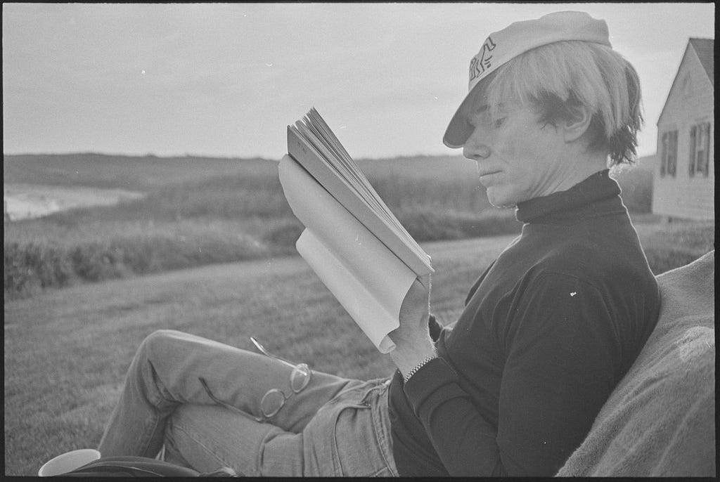 In a black and white photograph, Andy Warhol lounges on an outdoor chair while reading a book.
