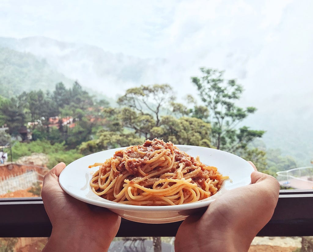 Two hands holding a bowl of cooked spaghetti with tomato sauce. A landscape of trees in the background.