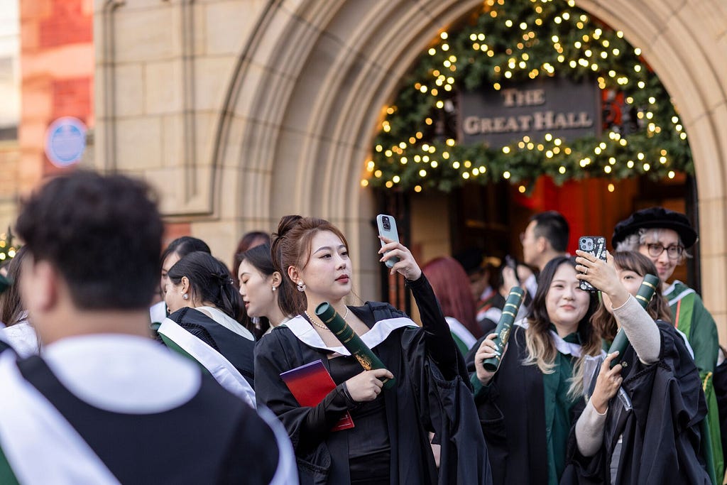 Graduates in suits and gowns take photos outside the Great Hall during the ceremony. Festive lights around the entrance enhance the event’s celebratory atmosphere.