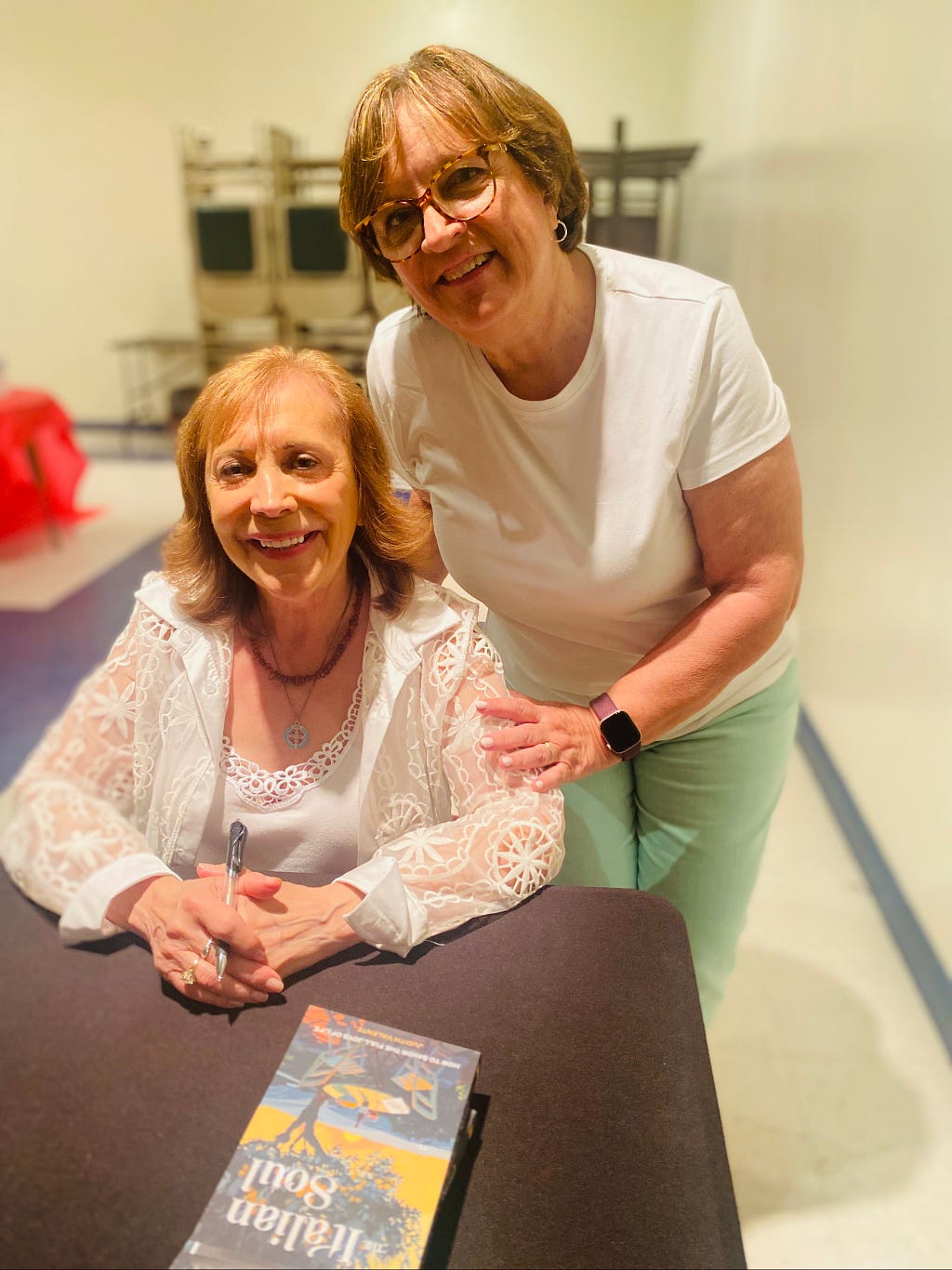 Author Judith Valente seated at a table with her book “The Italian Soul” and standing beside her is her elementary school classmate, Debby Stabulis.