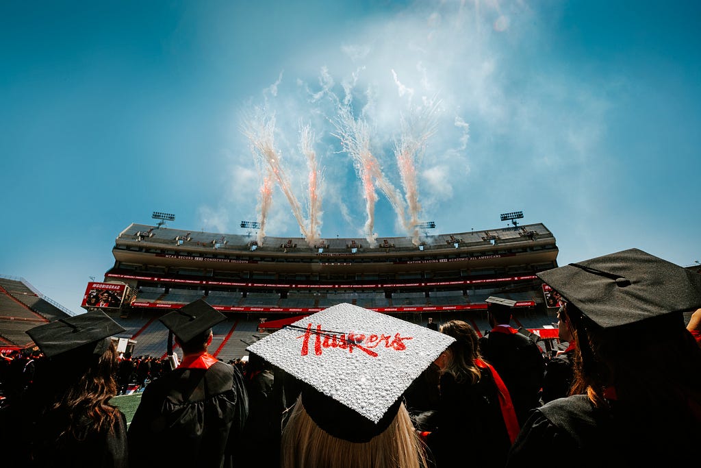 Fireworks in the shape of Ns erupt behind Memorial Stadium as graduates watch. A graduate whose mortarboard reads “Huskers” is centered in the foreground.