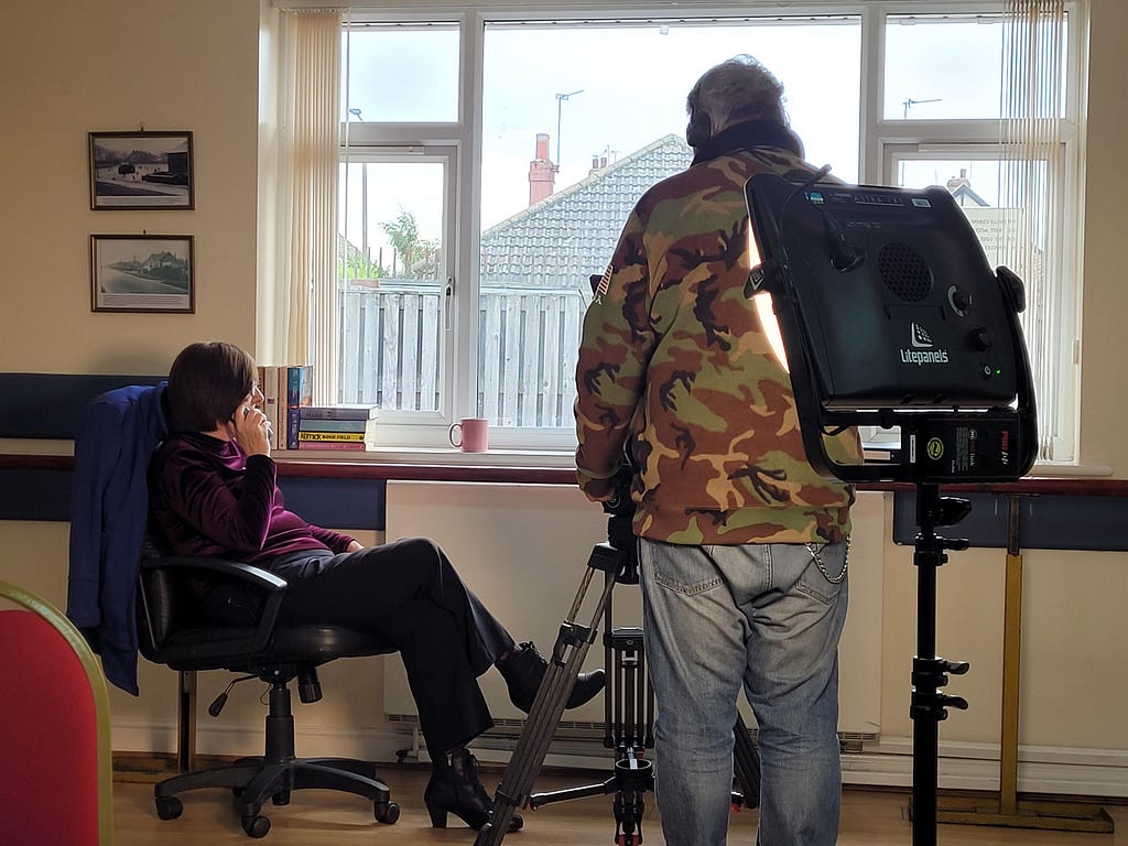 Fiona, an older woman, sits in a chair looking out the window while on the phone. A man films her with a TV camera.