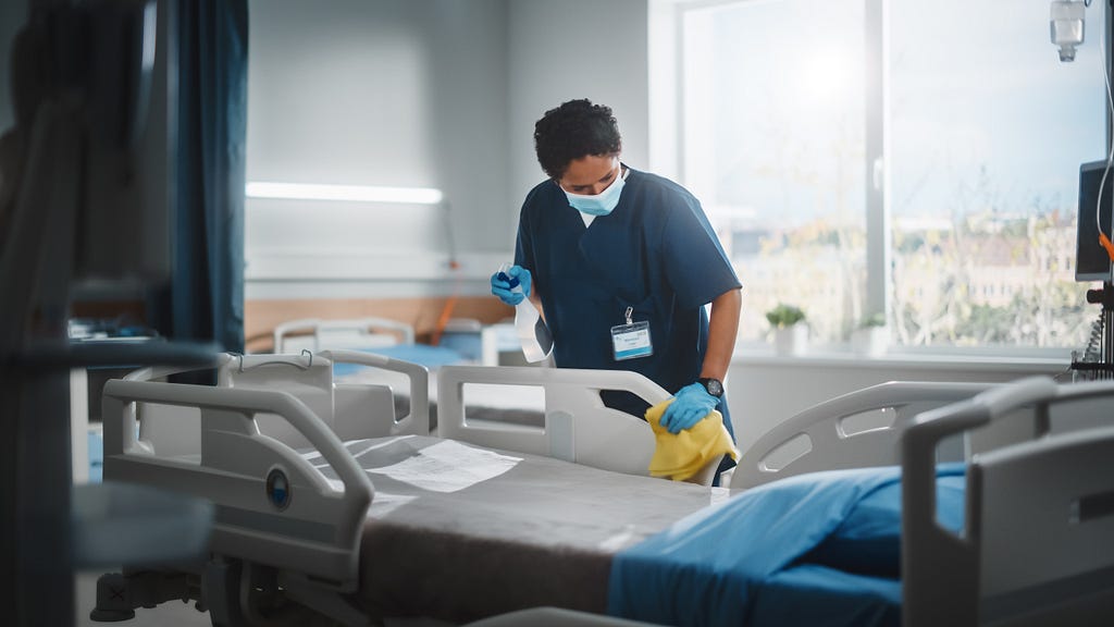 A masked and gloved health care worker disinfects a hospital bed.