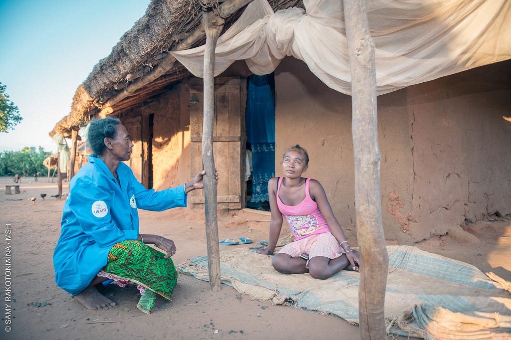 A community health volunteer speaks with a young woman sitting on the ground outside a mud building.