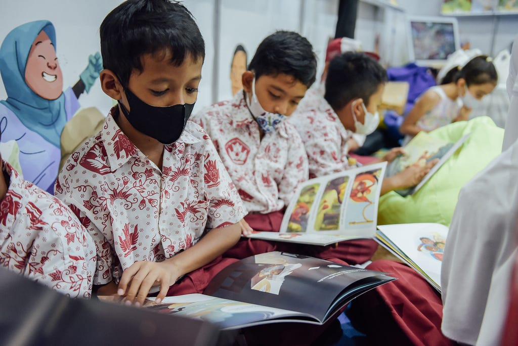 Three boys wearing matching white shirts with a red pattern and masks reading children’s books in a classroom setting.