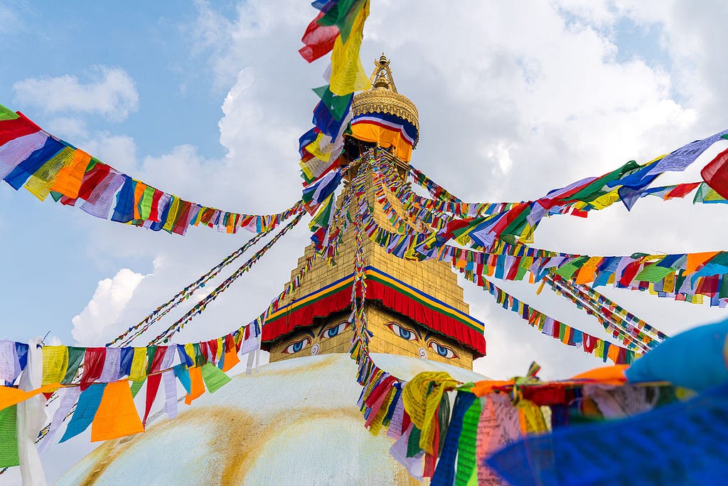 Colorful Buddhist prayer flags radiating from the golden spire of Boudhanath Stupa against a blue sky with wispy clouds.