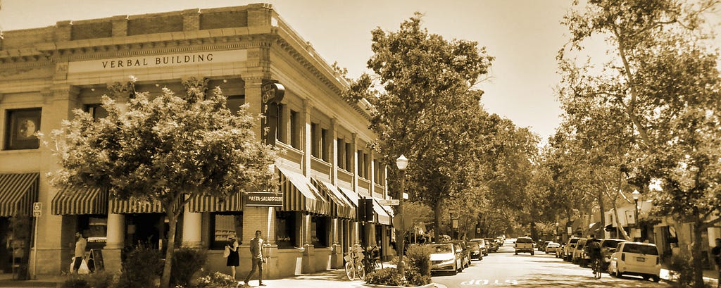 A street corner in Hunter Mudd College’s campus in Claremont, California. The “Verbal Building” is visible, as are pedestrians, pparked cars, and trees along the streeet.