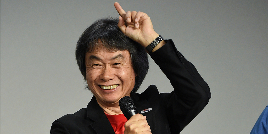 Shigeru Miyamoto on stage, pointing to the sky with a microphone in his hand.