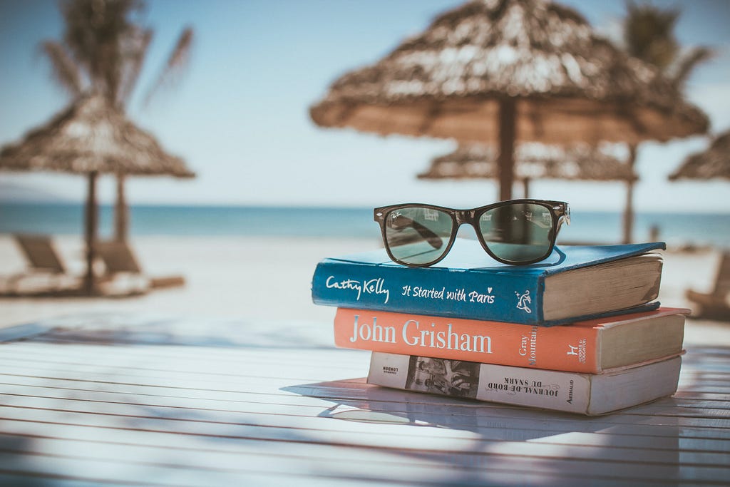 A sandy beach with clear skies. A stack of books and a pair of sunglasses sit beside lounge chairs and an umbrella.