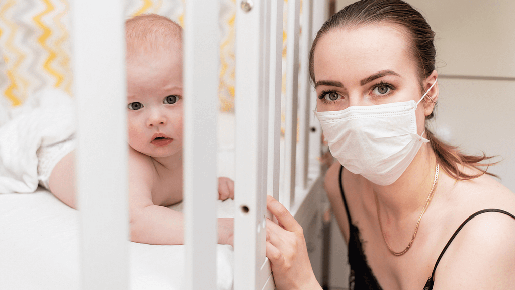 Baby looking through the bars of a cot, with his mom next to the cot wearing a mask.