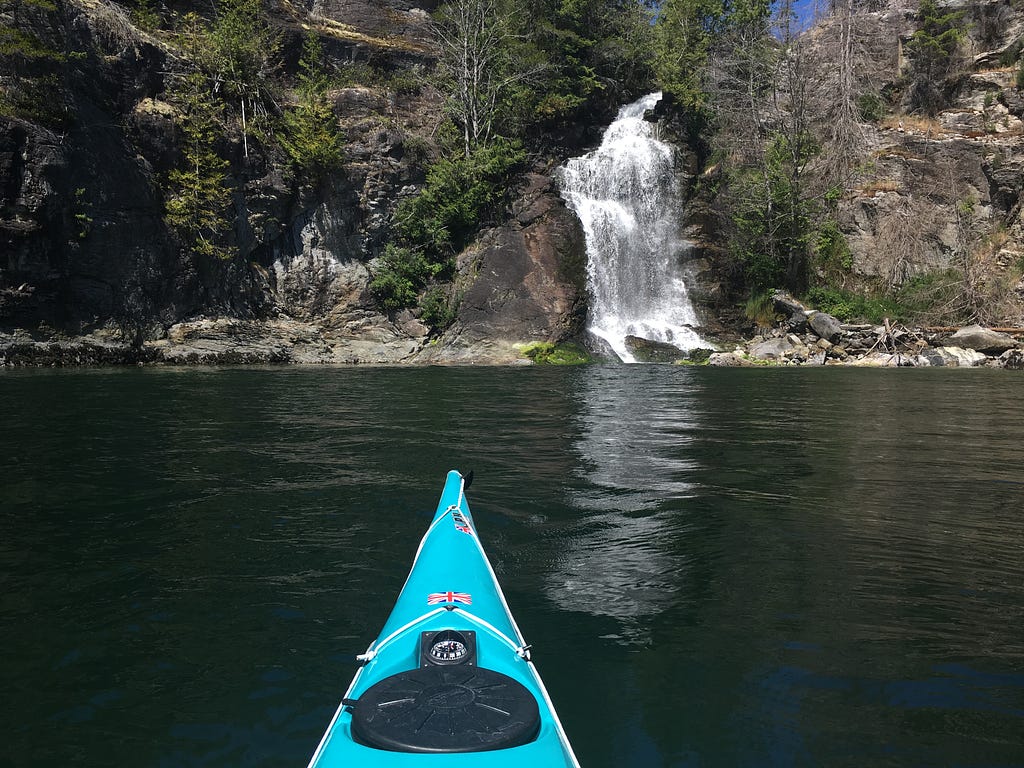 The nose of a kayak on the water, with a waterfall falling into the ocean