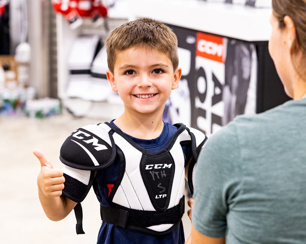 A young boy wearing hockey shoulder pads smiles and holds up a thumbs up. He is white with short brown hair. An adult woman with brown hair tied back in a ponytail and green shirt smiles at the boy with her back turned to the camera