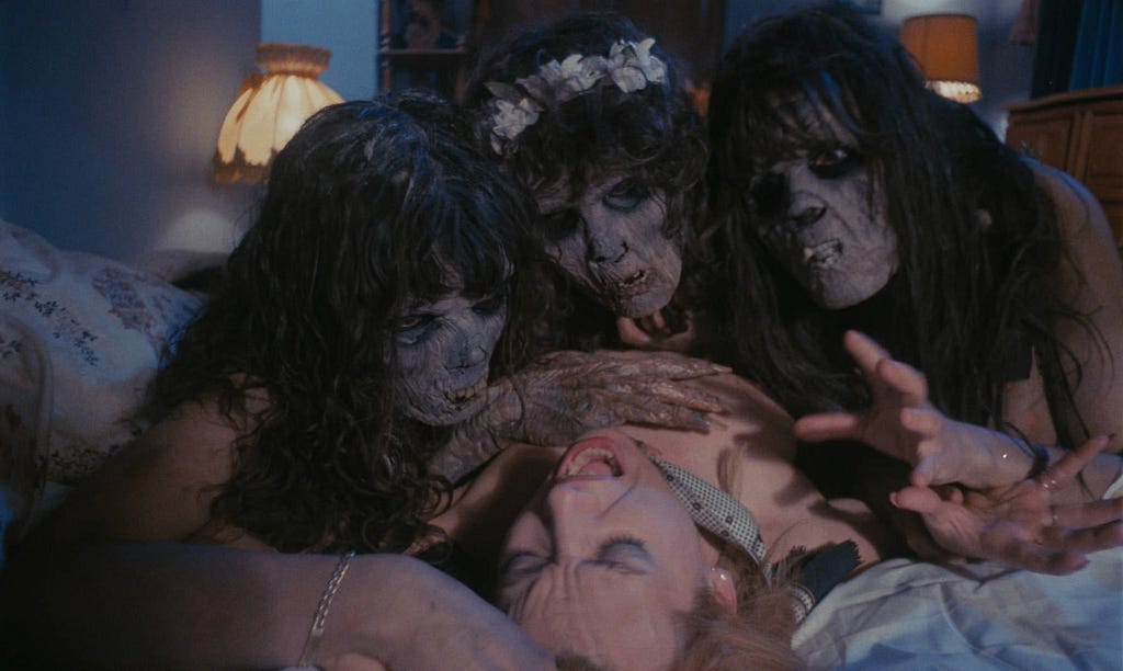 Three zombies women are seen taking old of a lone alive, nude woman laying on a bed.