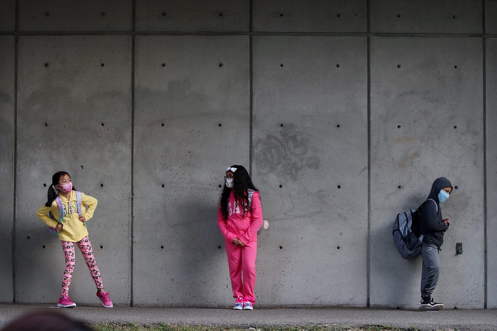 3 elementary school students wearing face masks and standing against a cement wall, 6 feet of distance between them.