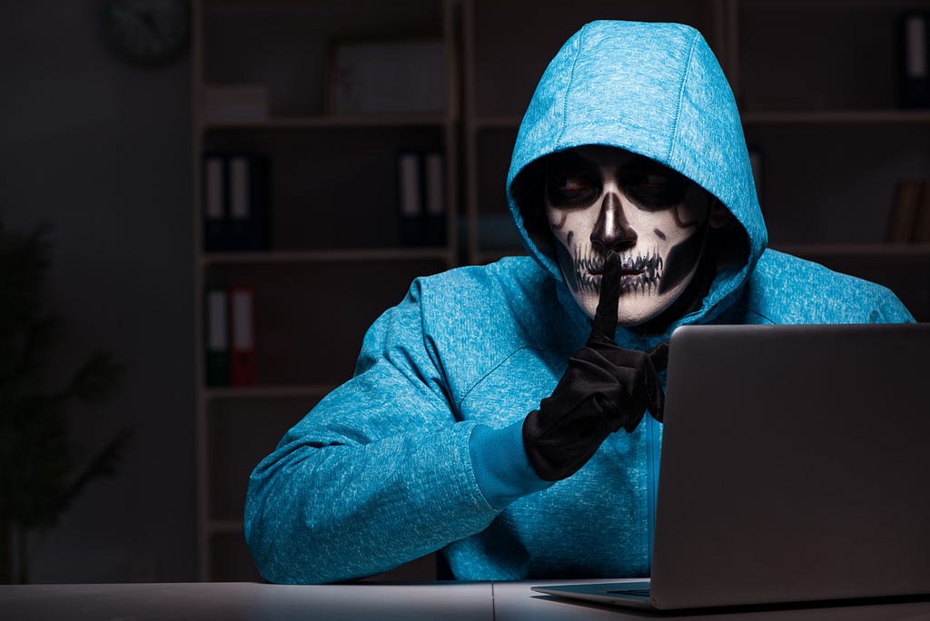 A person with skeleton face paint in a hoodie in a dark library behind a laptop, with their finger in a “shh” position.
