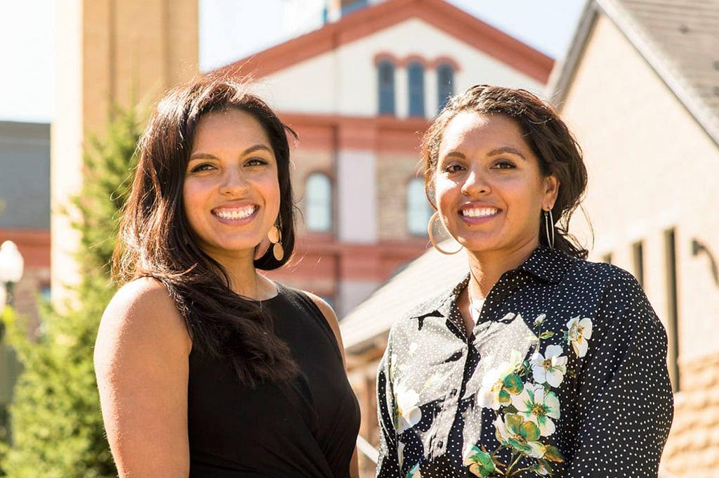 Diana and Denise Lopez stand side-by-side, smiling on the Regis Northwest Denver Campus with Main Hall and the Chapel in the background