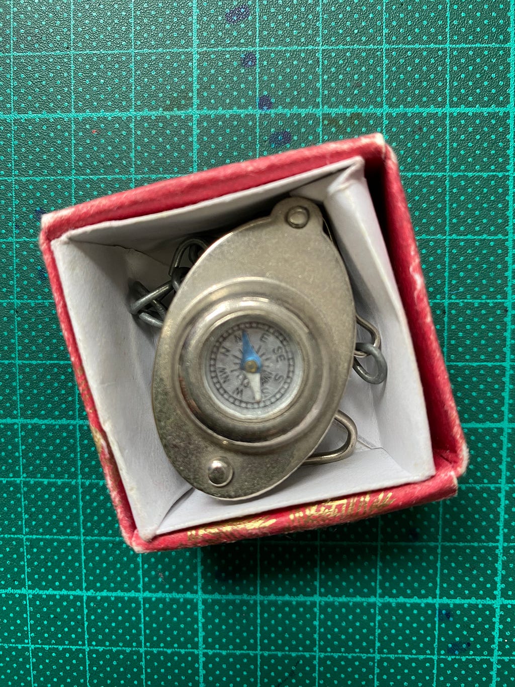 Picture of antique compass with stainless steel case and chain in small box. With green cutting mat as background.