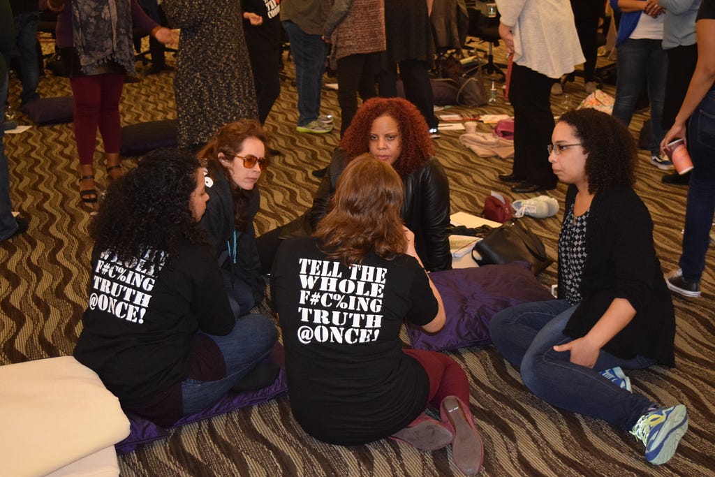 Photograph of five people sitting on the floor in a circle deep in conversation. Two people are wearing t-shirts with these words on the back: tell the whole f#c%ing truth @once!
