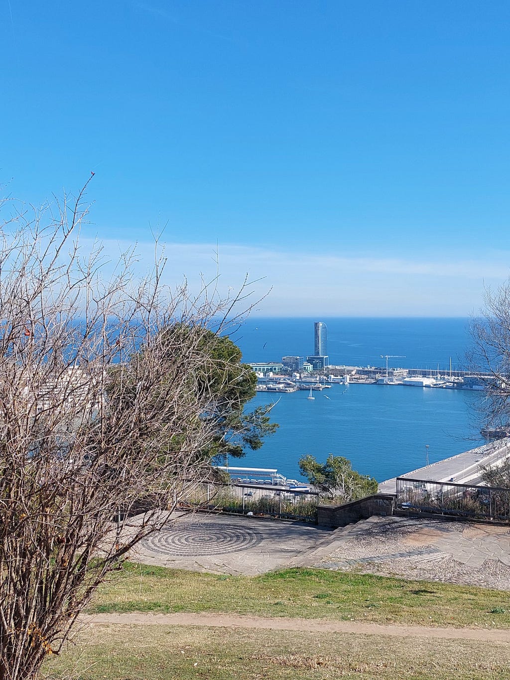 View of the port and the Mediterranean Sea from the Mirador Gardens, on the slope of Montjuic. In this image, you can see a sunny day, with the sea blending into the sky, some boats, the Hotel W, and some dry bushes due to winter.