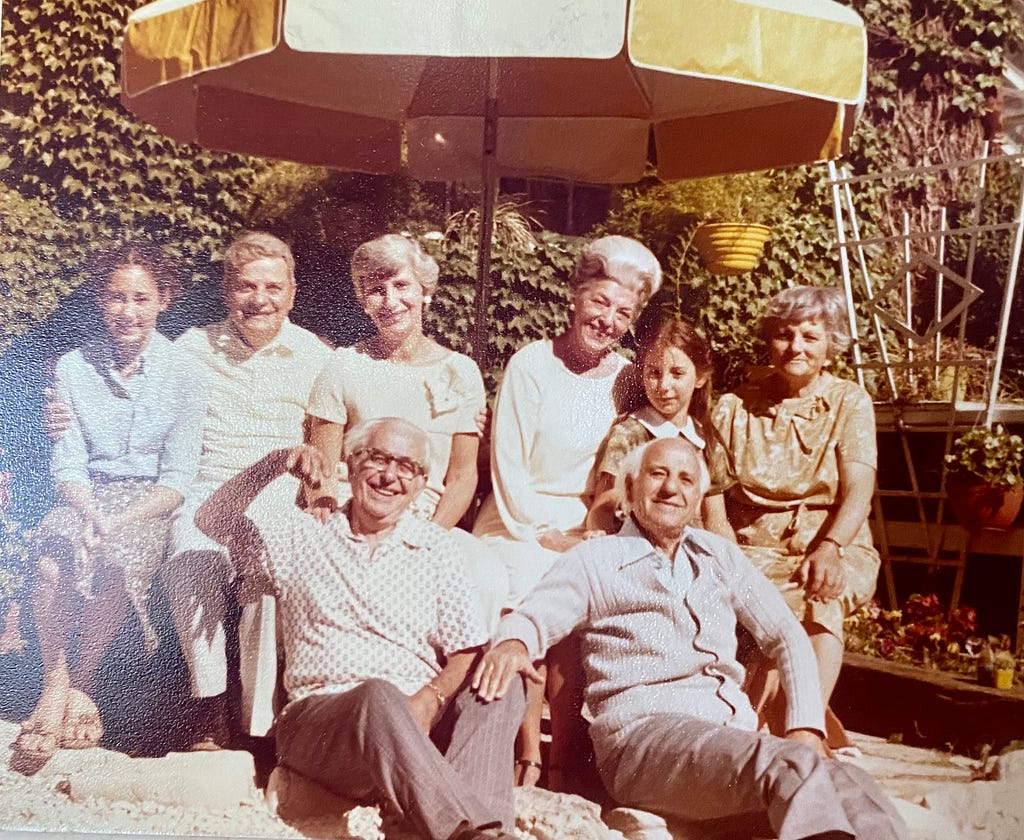 Smiling people seated under a picnic umbrella. 6 in the back row, 2 seated on the ground in the front row.