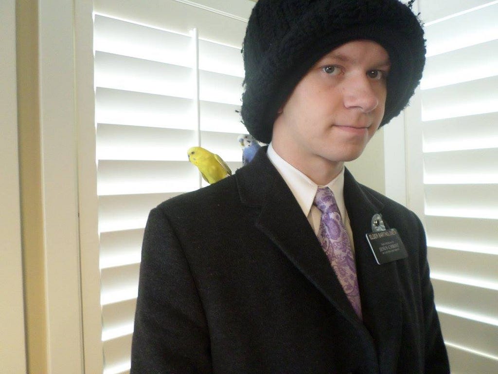 A young man wearing a knit hat on a mormon mission, with a couple small pet birds on his shoulder.