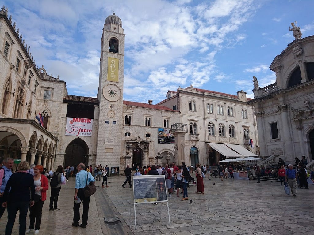A bell tower in the old town, an old church on the right with tourists and locals spending leisurley time