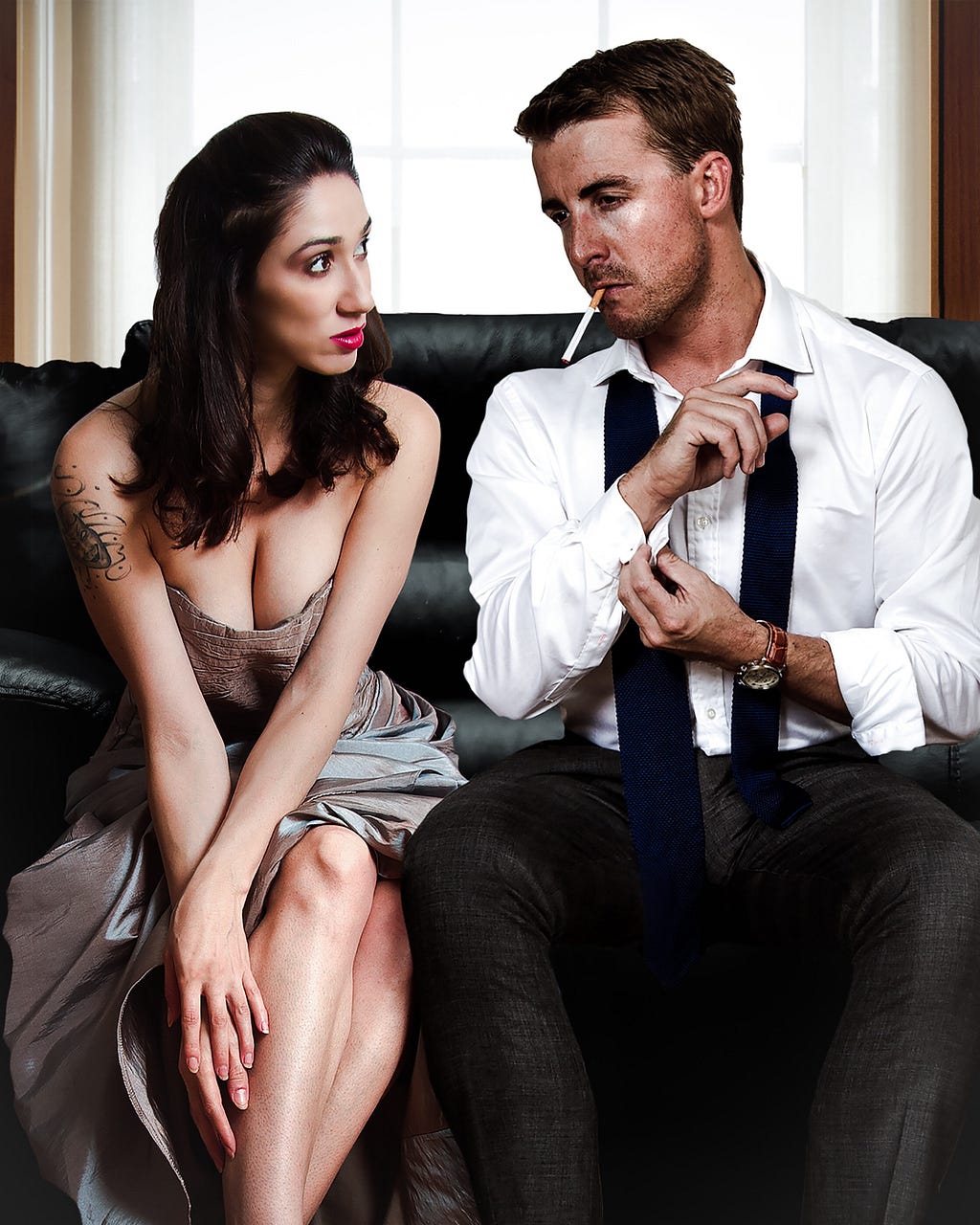 A white woman and man wearing formal attire, looking at each other, and sitting on a couch.