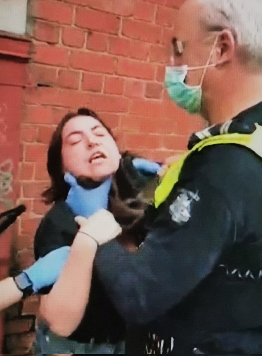 A short woman, eyes closed, grabbing at the wrist of a grey-haired cop with gloved hands around her neck, and upper arm held by another gloved hand which is only visible in-frame to the forearm