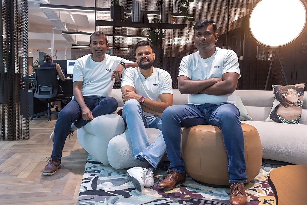Three middle-aged south asian men sitting casually on brown and cream office furniture, dressed in jeans and white t-shirts with the Hola Health logo.