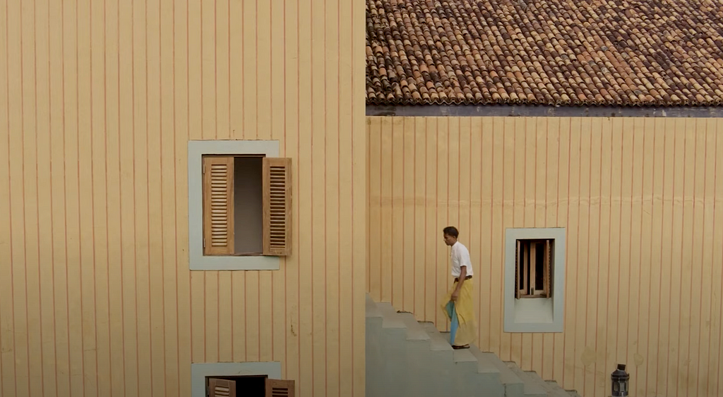 Staff member of the Lighthouse hotel walking up a staircase outside the building. Walls of the building is painted in a similar color to the sand and with wooden windows. Staff member is wearing a similar coloured sarong with accent of light blue matched with a white shirt.