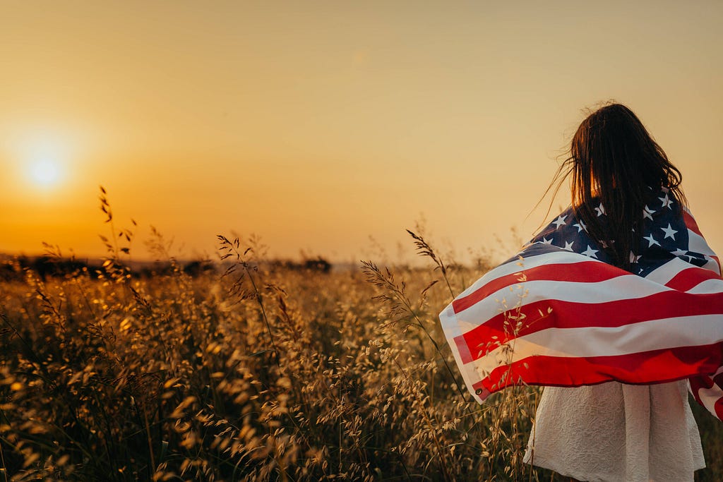 Young girl with American flag wrapped around her shoulders looks out at wheat field at sunset.