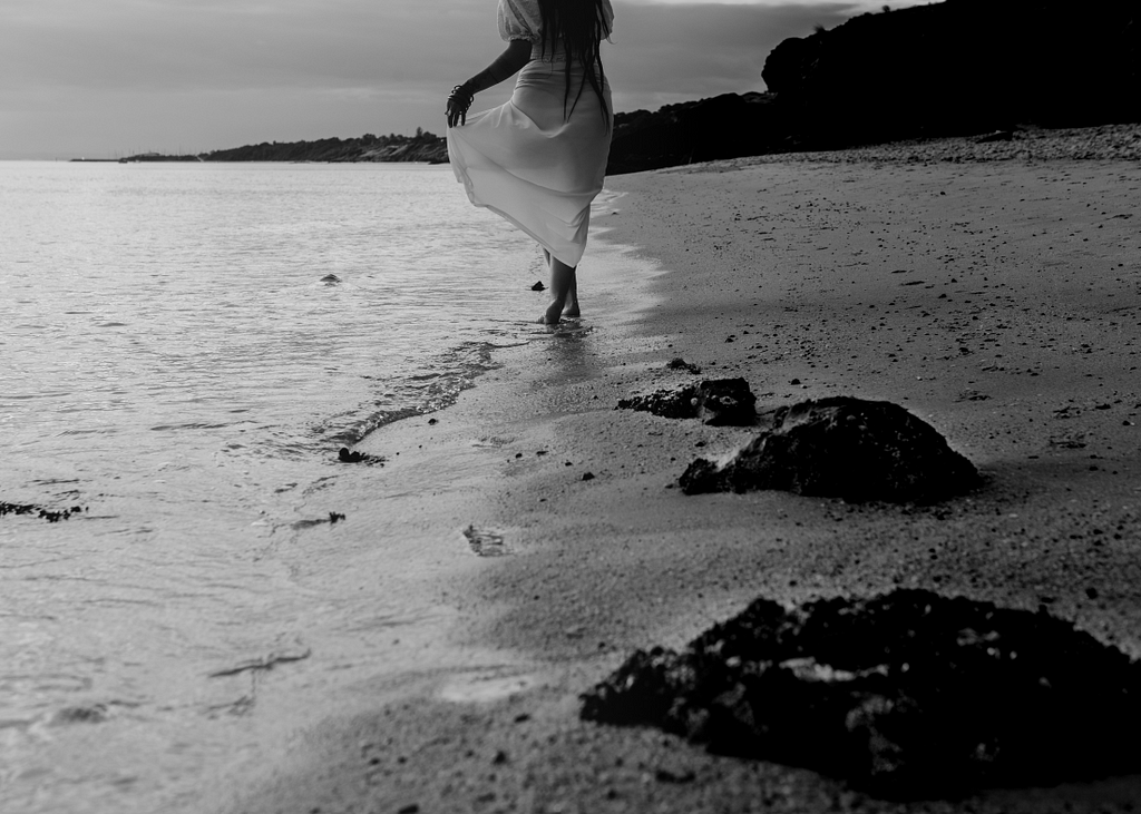 A woman in a long, white flowing dress walks leisurely along an empty beach. Her feet are lightly touched by the incoming waves.