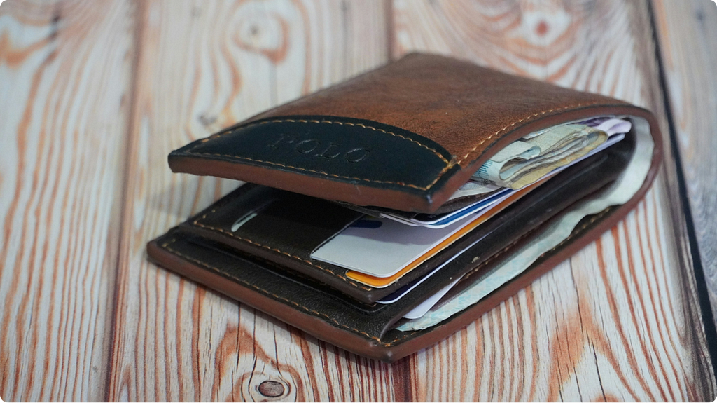 A brown leather wallet on a wooden table, slightly open, with several debit and credit cards visible alongside folded banknotes.